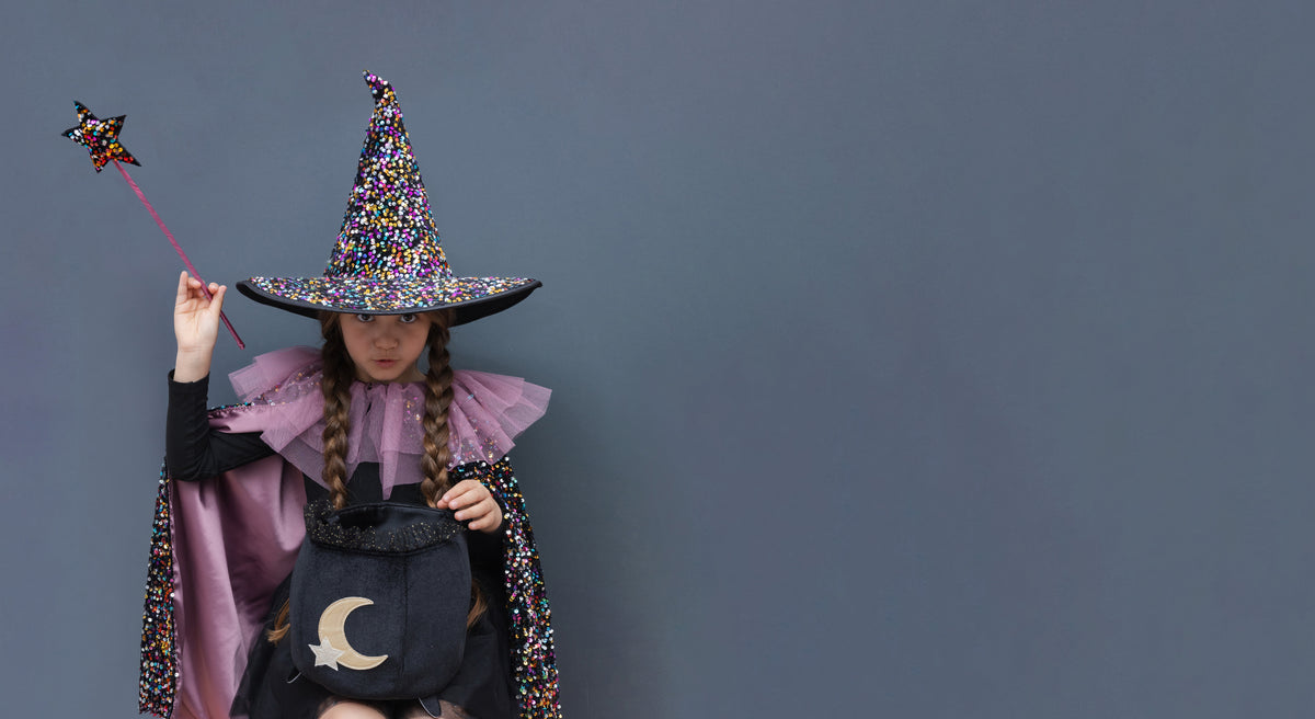 A child wearing a sparkly multicolor sequinned witch costume with a pointed hat, cape with purple ruffled collar, and a black cauldron bag with a gold crescent moon, holding a star wand against a solid dark gray background.