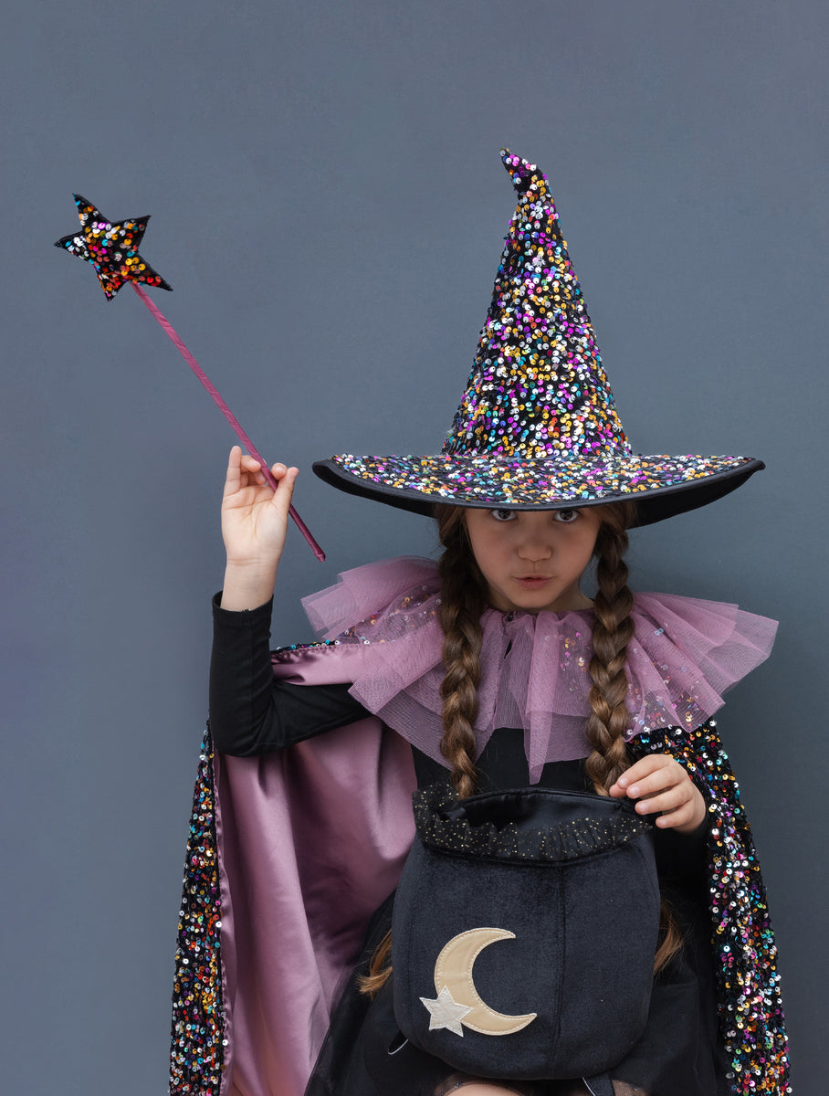 A child wearing a sparkly multicolor sequinned witch costume with a pointed hat, cape with purple ruffled collar, and a black cauldron bag with a gold crescent moon, holding a star wand against a solid dark gray background.